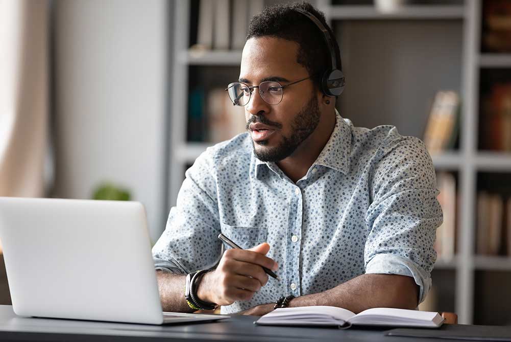 a man sits at a laptop computer with a headset on, holding a pen over a notebook and engaged in conversation